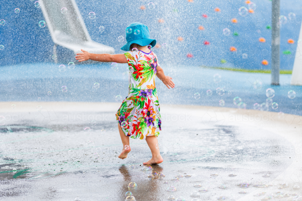 Young child running barefoot through water fountain play area at park in swim hat and dress - Australian Stock Image