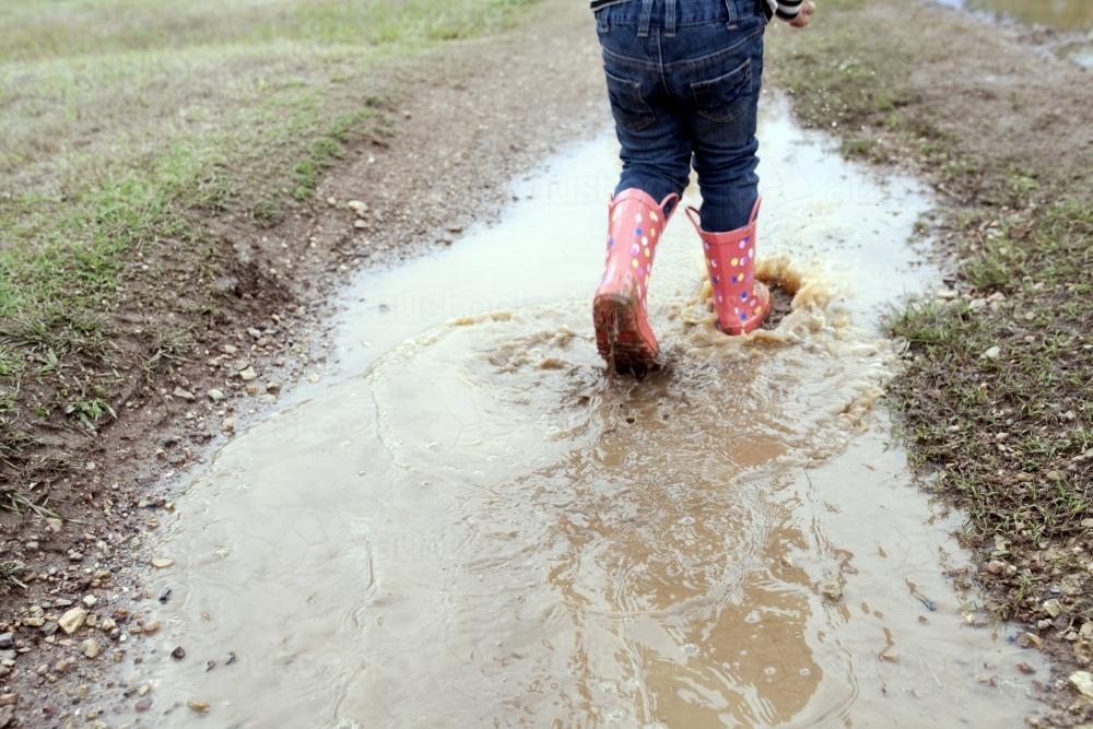 Young child wearing gumboots splashing through muddy puddle - Australian Stock Image
