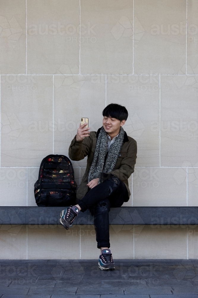 Young Chinese man sitting on bench using mobile phone - Australian Stock Image