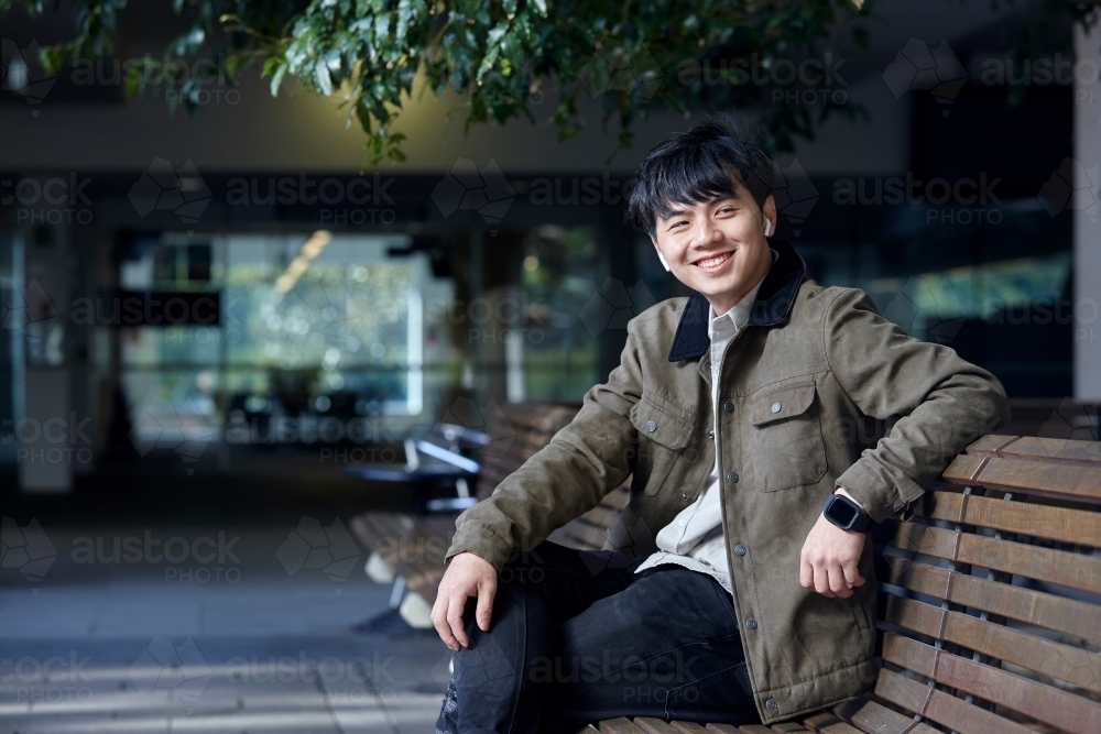 Young Chinese man smiling whilst listening to wireless headphones - Australian Stock Image
