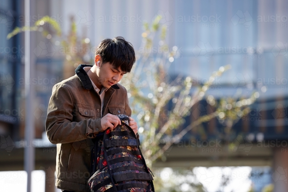 Young Chinese student outdoors opening backpack - Australian Stock Image