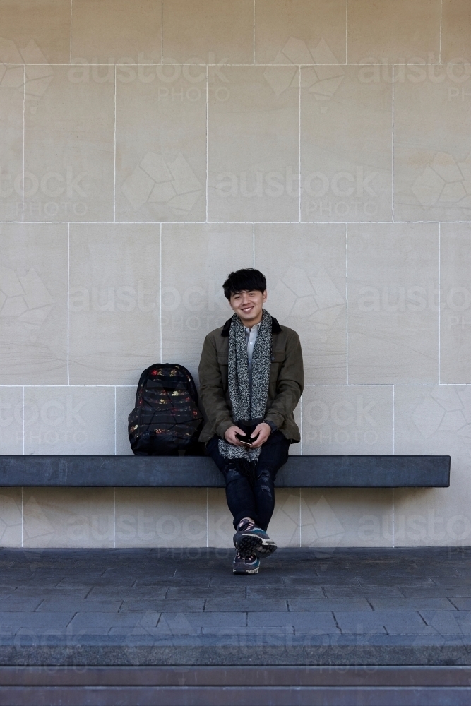 Young Chinese student sitting on bench at university campus with mobile phone - Australian Stock Image