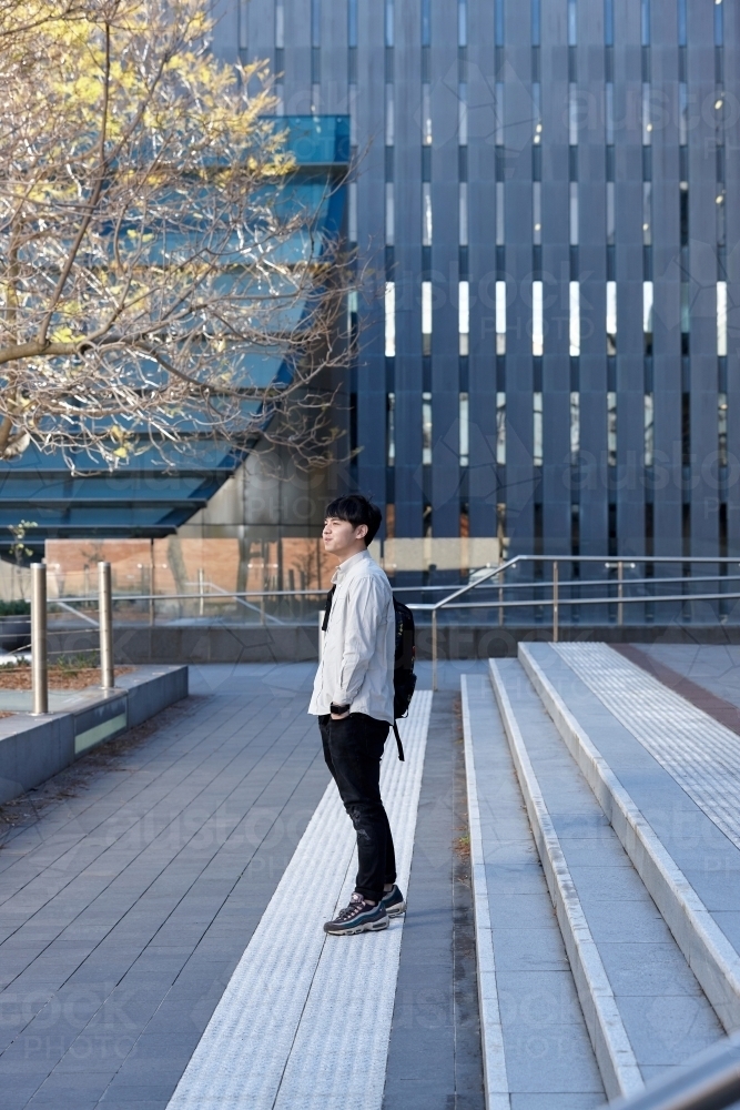 Young Chinese student standing on stairs smiling with buildings and tree behind - Australian Stock Image
