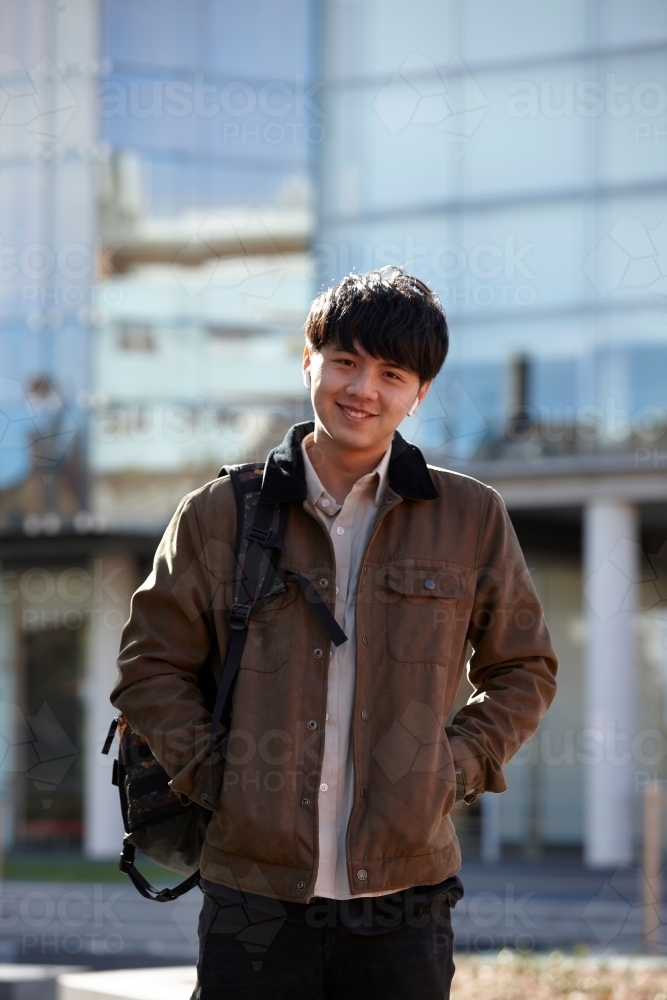 Young Chinese student standing with hands in pocket with listening to wireless headphones : Austockphoto Young Chinese student standing with hands in pocket with listening to wireless headphones - Australian Stock Image