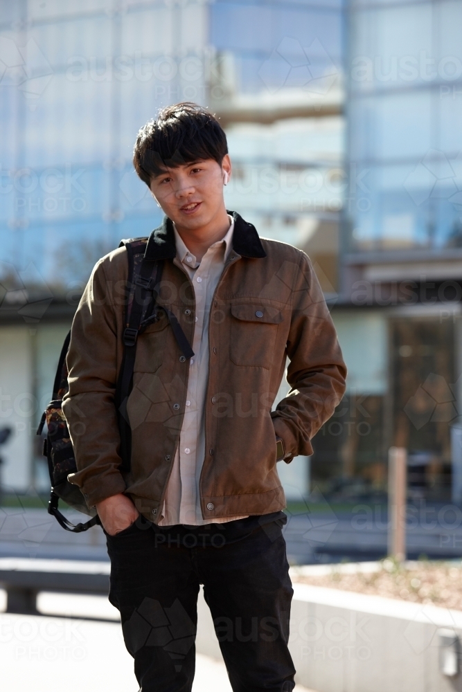 Young Chinese student standing with hands in pocket with listening to wireless headphones - Australian Stock Image