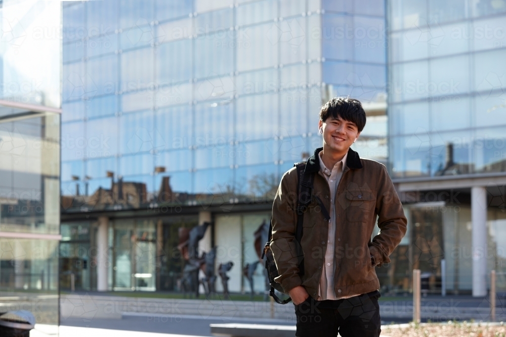 Young Chinese student standing with hands in pocket with listening to wireless headphones - Australian Stock Image
