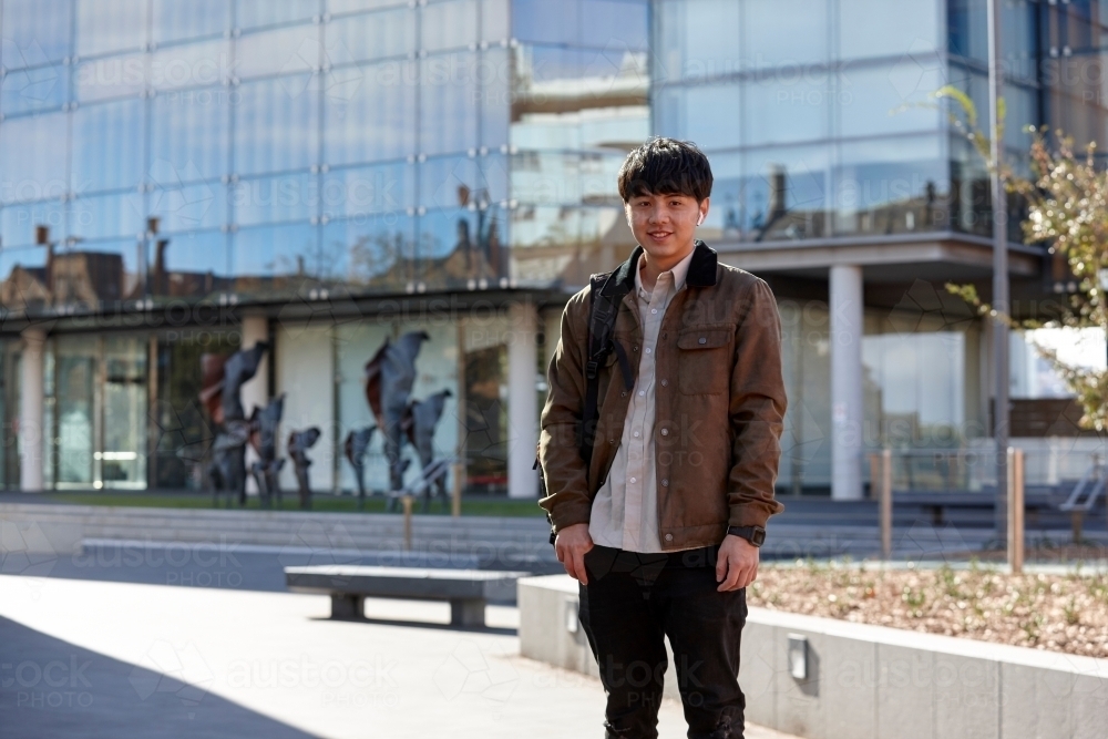 Young Chinese student standing with hands in pocket with listening to wireless headphones - Australian Stock Image