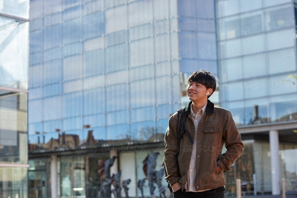 Young Chinese student standing with hands in pocket with listening to wireless headphones - Australian Stock Image