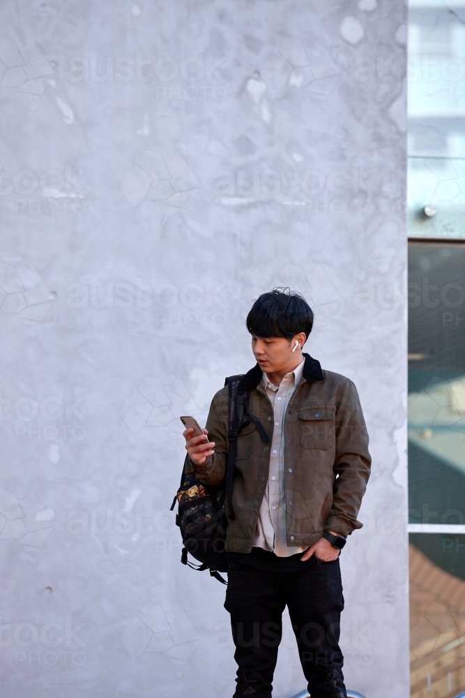 Young Chinese university student standing using mobile phone and wireless headphones - Australian Stock Image