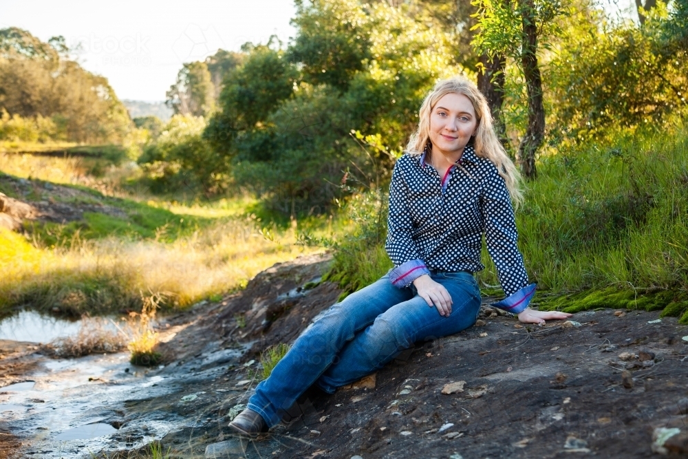 Young country woman sitting on stones beside creek - Australian Stock Image
