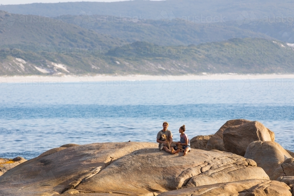 young couple and dog sitting on granite rock by the sea - Australian Stock Image