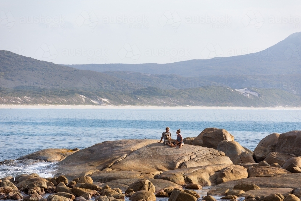 young couple and dog sitting on granite rock by the sea - Australian Stock Image