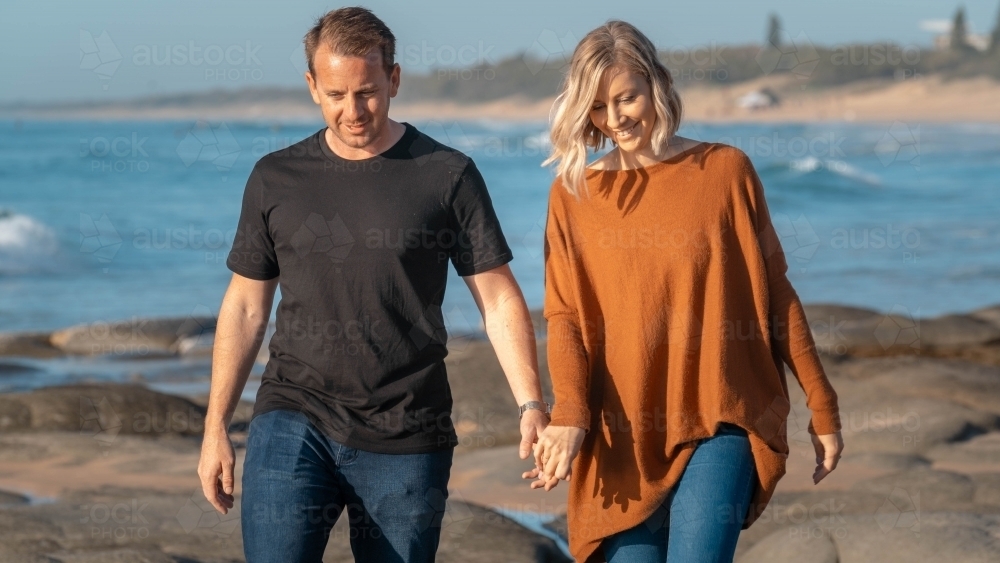 Young couple holding hands walking on rocks at beach - Australian Stock Image