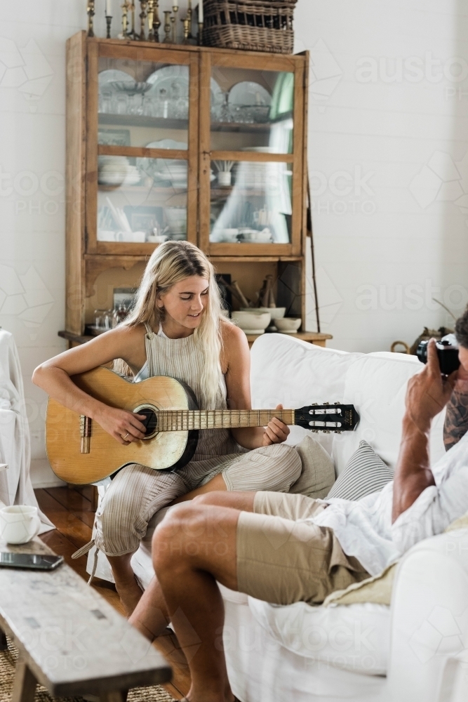 Young couple sitting on a couch, with man taking  a photo of woman playing guitar. - Australian Stock Image