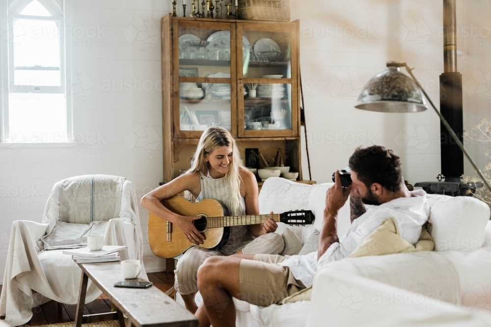 Young couple sitting on a couch, with man taking photo of  woman playing guitar. - Australian Stock Image