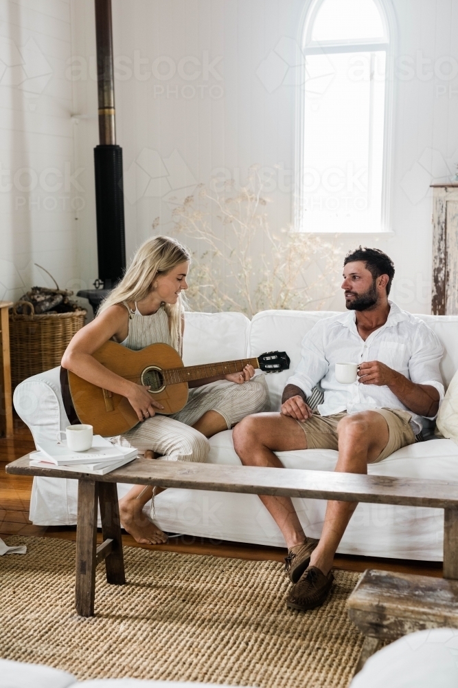 Young couple sitting on a couch, with the woman playing guitar. - Australian Stock Image