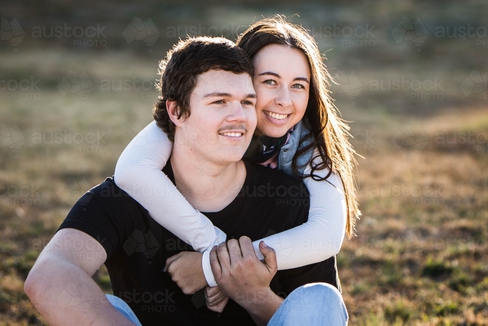 Young couple sitting on grass woman's arms around mans neck smiling - Australian Stock Image