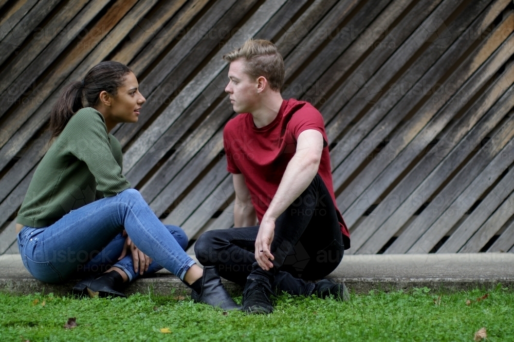 Young couple sitting on the ground talking in front of textured wooden panels - Australian Stock Image