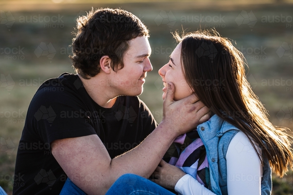 Young couple sitting together laughing with man holding girlfriend's face - Australian Stock Image