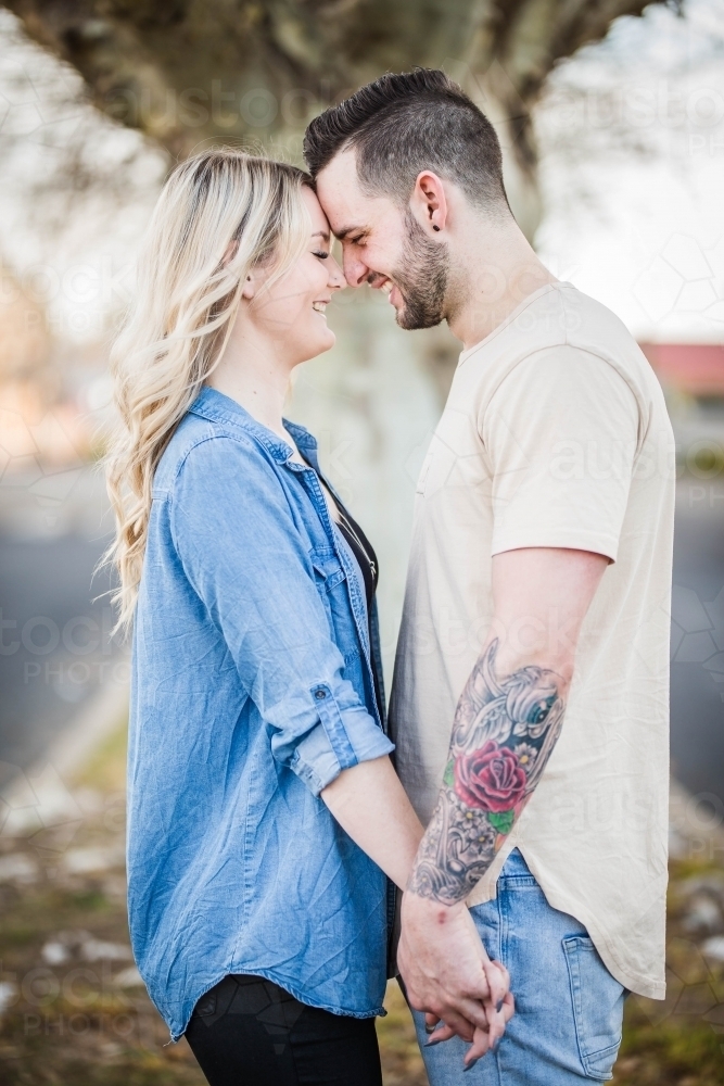 Young couple standing close together holding hands foreheads touching laughing - Australian Stock Image
