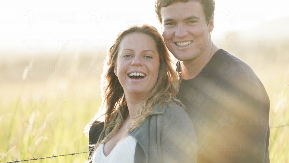 Young couple standing in field, looking at camera, sun flare behind - Australian Stock Image