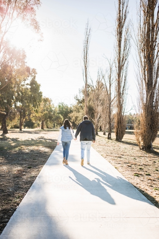 Young couple walking away into distance on path in park holding hands - Australian Stock Image