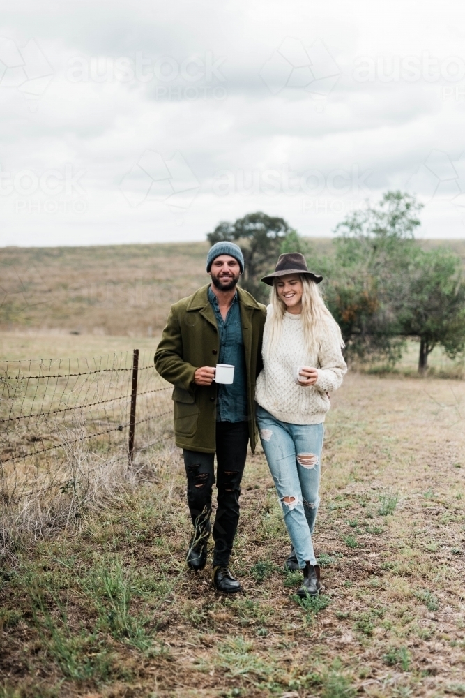 Young couple walking through farm holding cups of coffee - Australian Stock Image