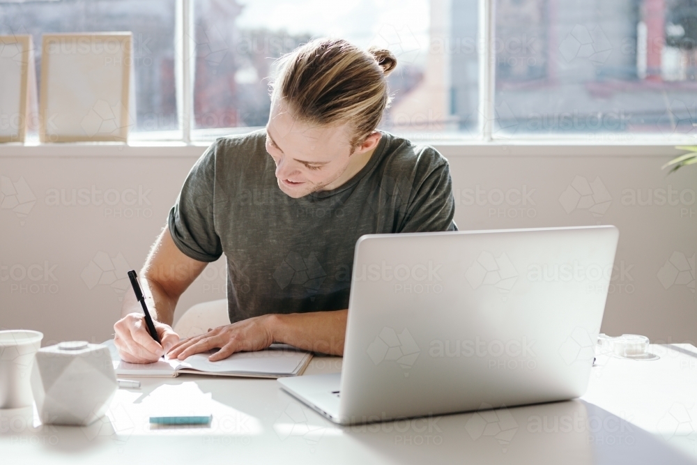 Young creative guy with blond bun making notes in a studio - Australian Stock Image