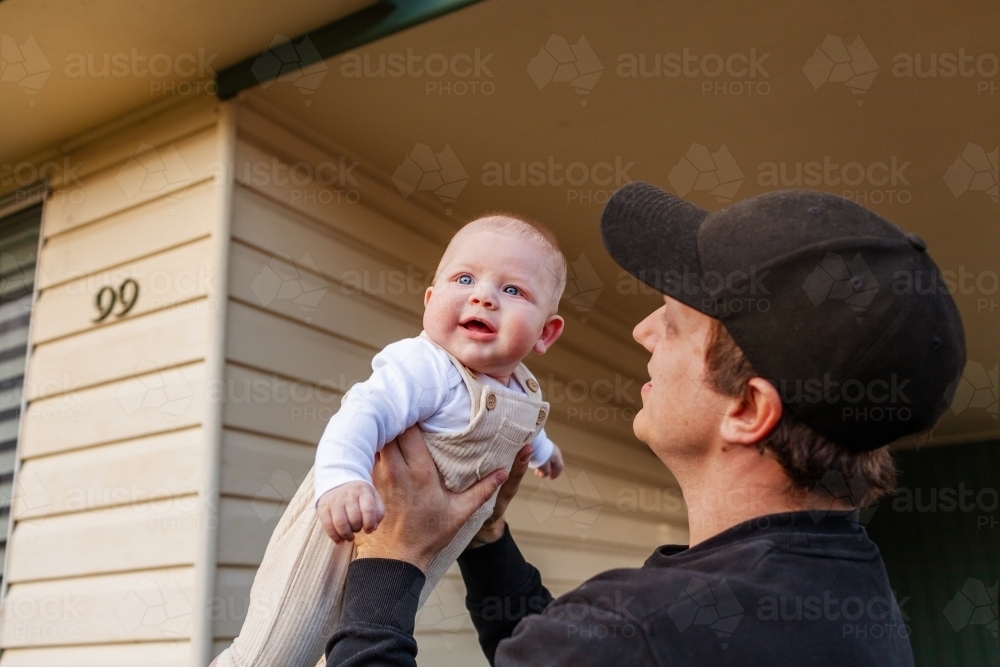 Young dad holding his baby son up in the air outside family home - Australian Stock Image