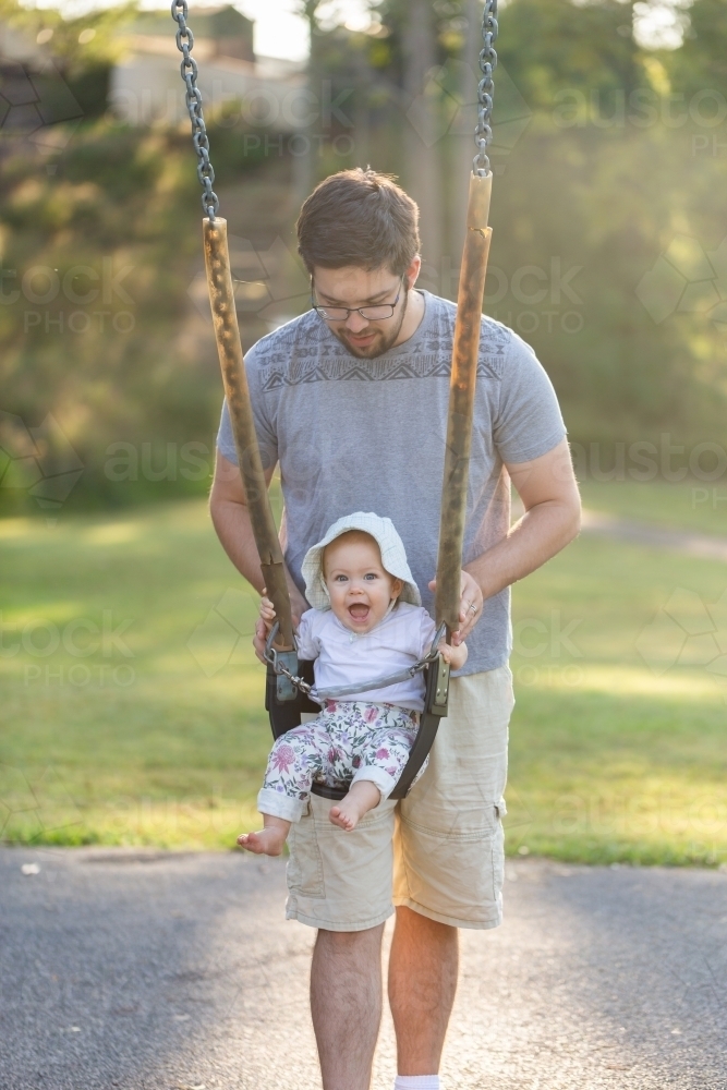 Young dad pushing baby girl on swing at park in morning - Australian Stock Image