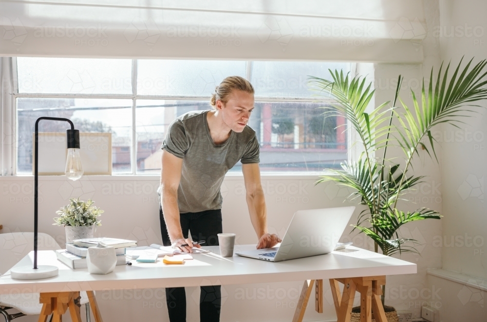 Young designer standing at a desk looking at a laptop - Australian Stock Image