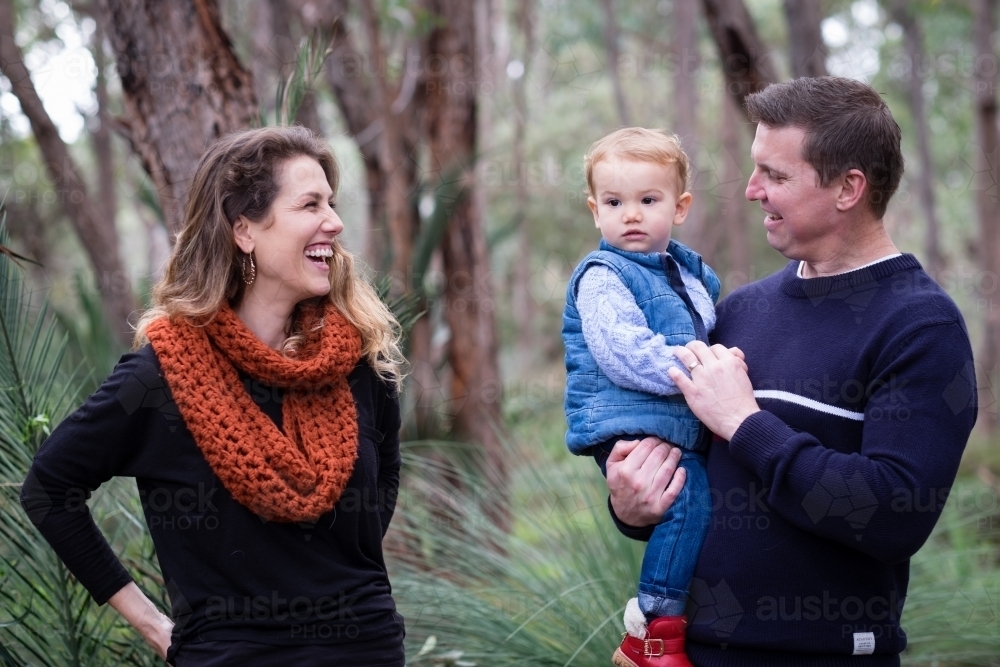 Young family portrait in the bush, mother laughing and  looking onto father holding toddler smiling - Australian Stock Image