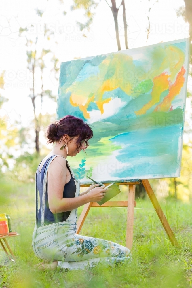 young female artist working on green landscape painting outdoors seen through grass - Australian Stock Image