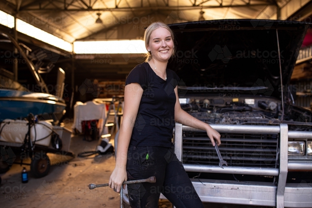 Young female aussie mechanic holding tools and leaning on a car in need of repair in workshop garage - Australian Stock Image