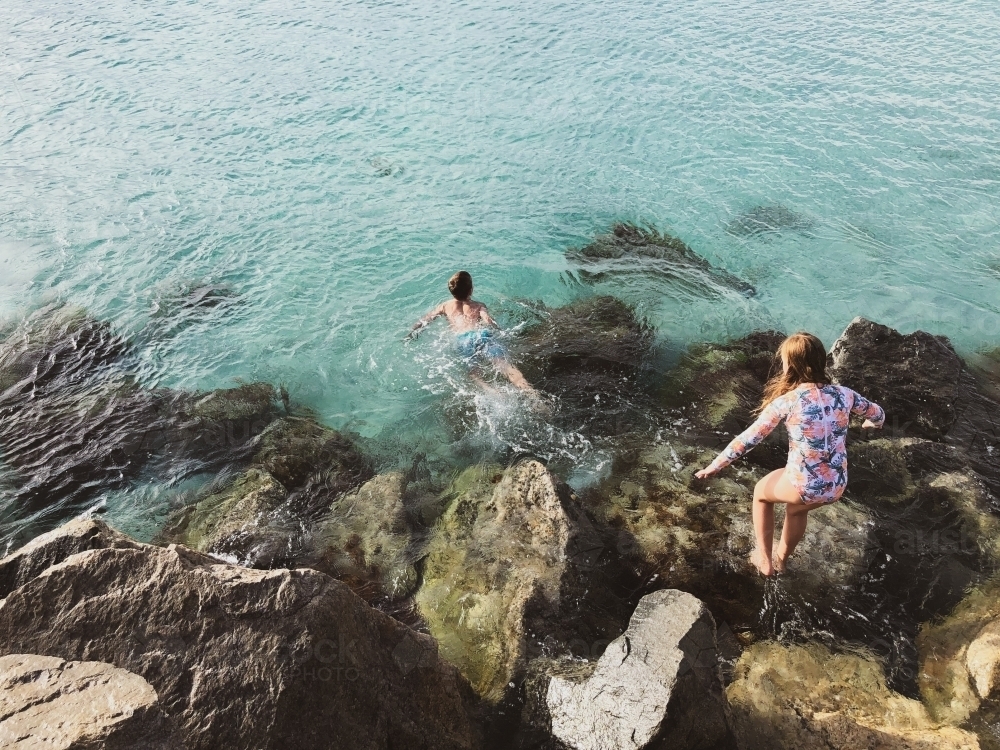 Young girl and boy climbing on coastal rocks and jumping into ocean - Australian Stock Image