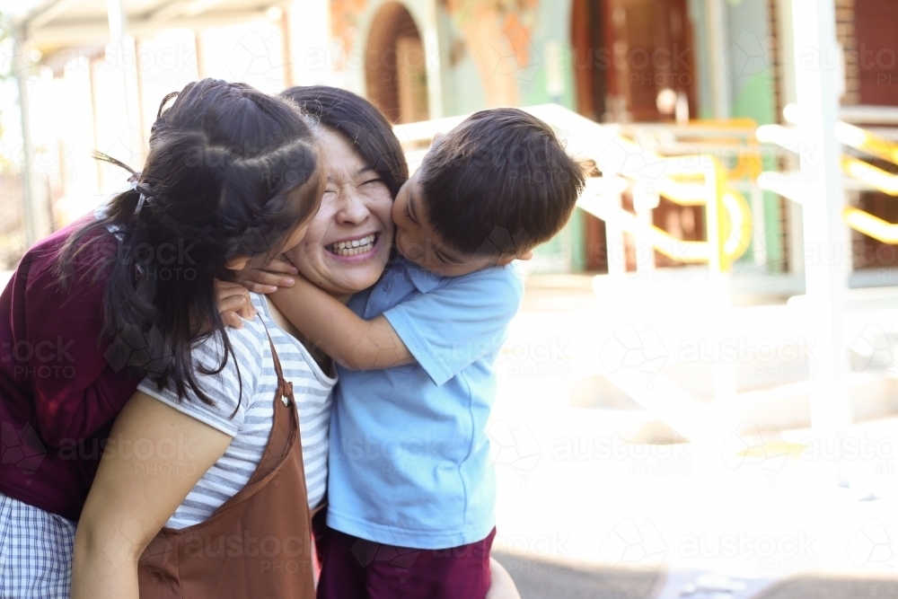 Young girl and boy in school uniform hugging and kissing their mum at school - Australian Stock Image