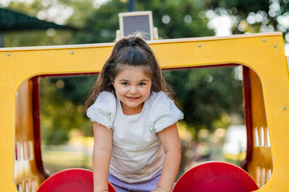 Young girl climbs on yellow play structure in a park while smiling - Australian Stock Image