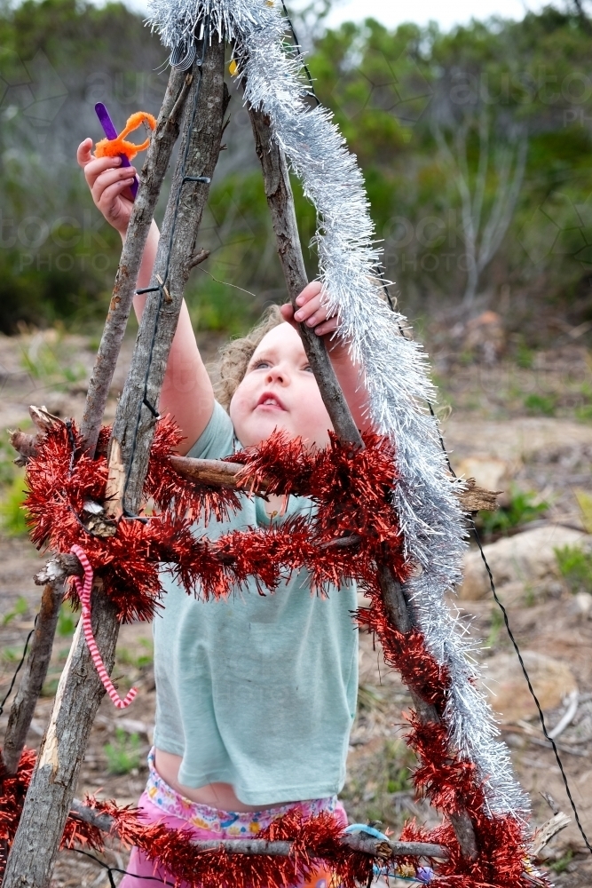 Young girl decorating Bush Christmas tree with tinsel in rural location. - Australian Stock Image