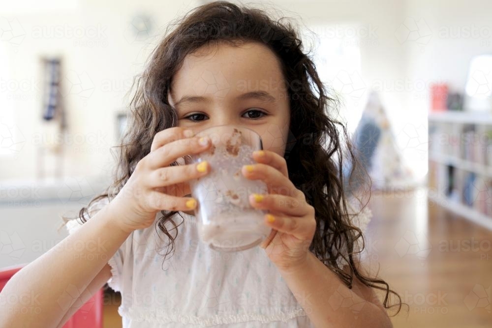 Young girl drinking chocolate milk smiling behind glass - Australian Stock Image