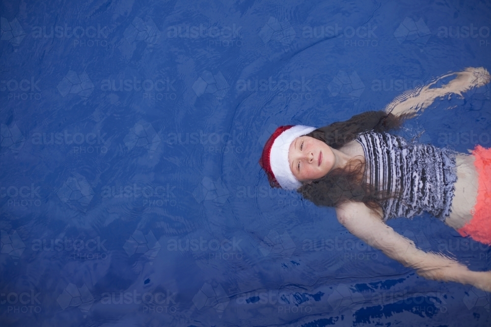 Young girl floats in a pool wearing a festive christmas santa hat - Australian Stock Image