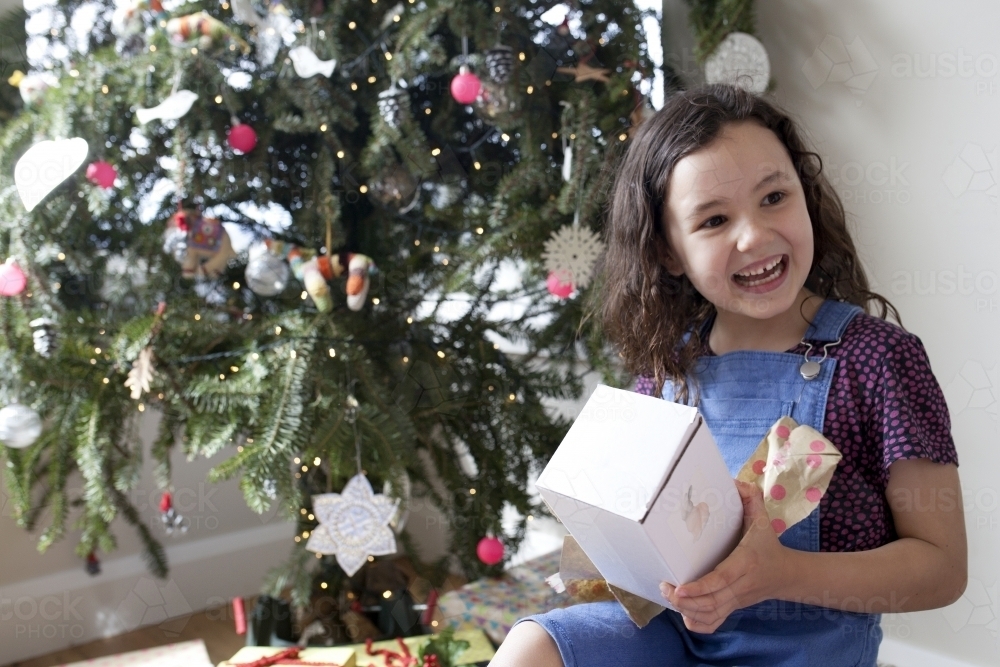 Young girl happy and smiling after opening gift in front of Christmas tree - Australian Stock Image
