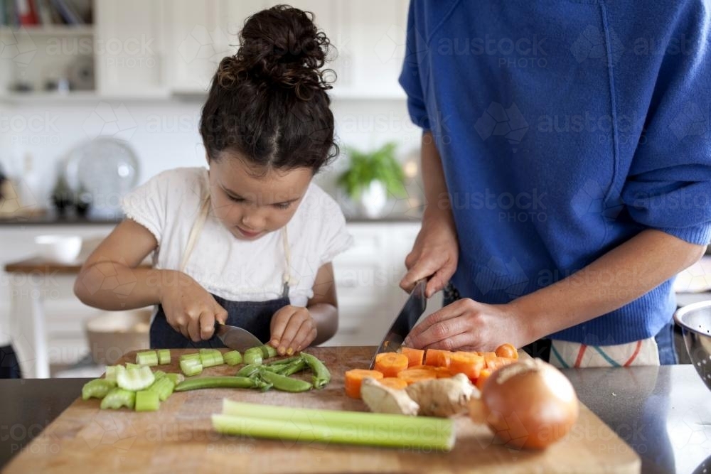 Young girl helping cut vegetables with mother in kitchen - Australian Stock Image