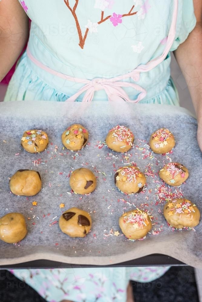 young girl holding a tray of uncooked cookies, covered with sprinkles - Australian Stock Image
