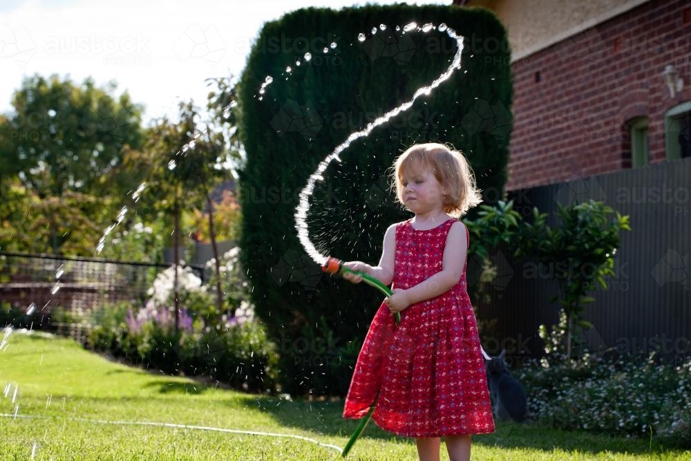 Young girl in a red dress playing with water and a hose in backyard - Australian Stock Image