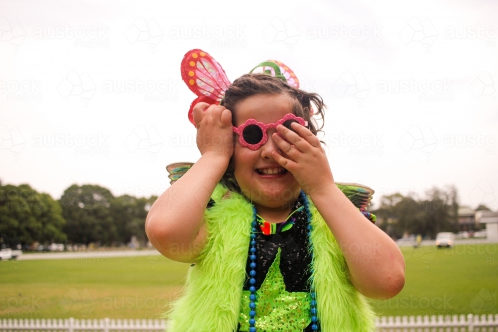 Young girl in fluoro dress-up covering one eye of pink glasses - Australian Stock Image