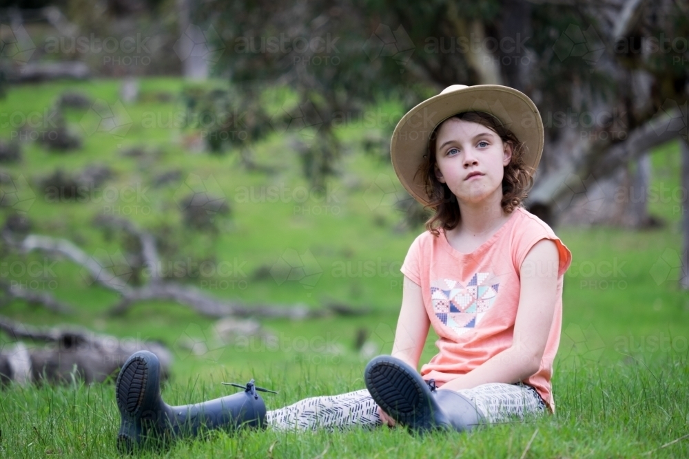 Young girl in gumboots and akubra sitting in open field - Australian Stock Image