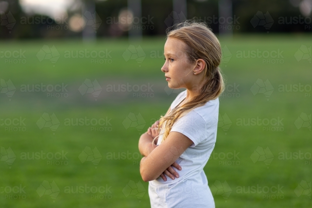 young girl in profile standing with arms crossed outside on playing field - Australian Stock Image
