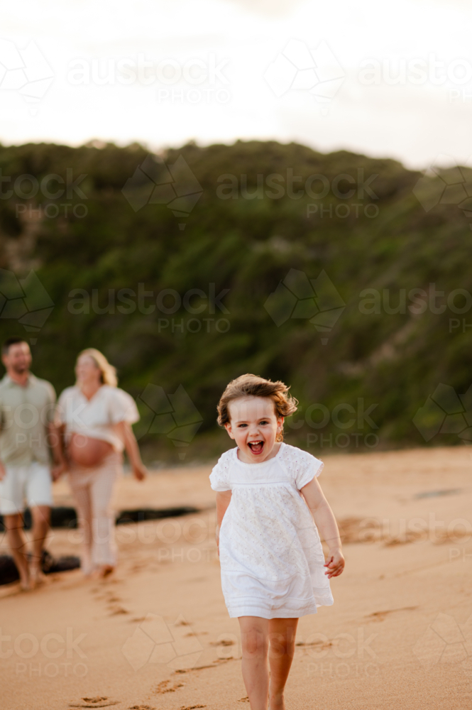 Young girl in white dress runs excitedly on sandy beach while family walks behind her at sunset - Australian Stock Image