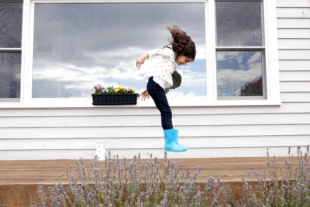 Young girl jumping on wooden deck in blue gumboots - Australian Stock Image