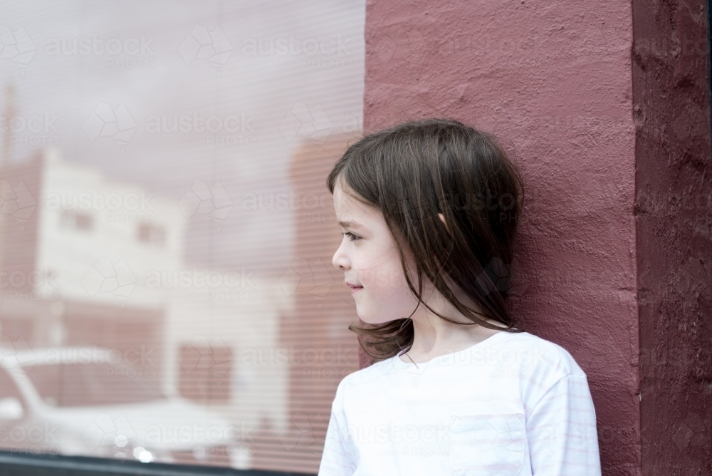 Young girl looking away, leaning against a pink brick wall with a window reflecting the street - Australian Stock Image